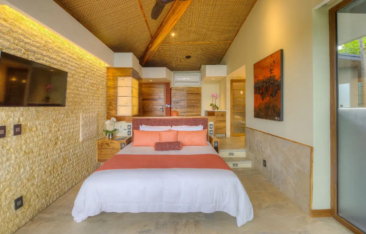 Tropical modern bedroom design featuring a king bed with terracotta bedding, rough stone accent wall, and woven ceiling