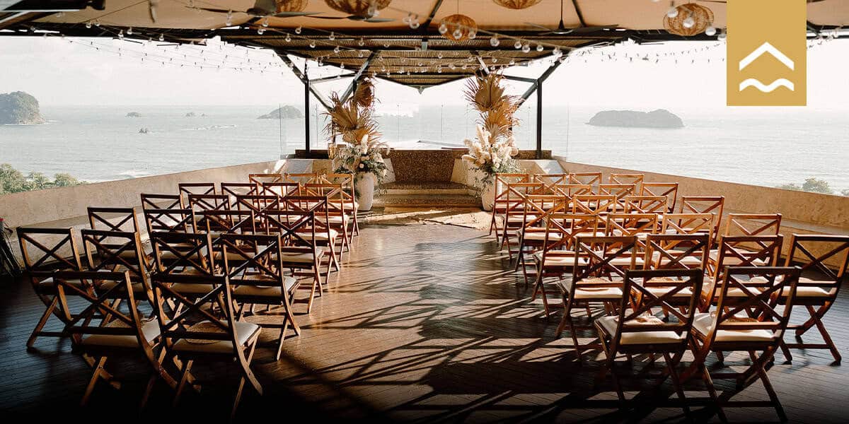Elegant oceanview wedding ceremony setup with wooden chairs, floral arrangements, string lights, and panoramic backdrop of islands and sea