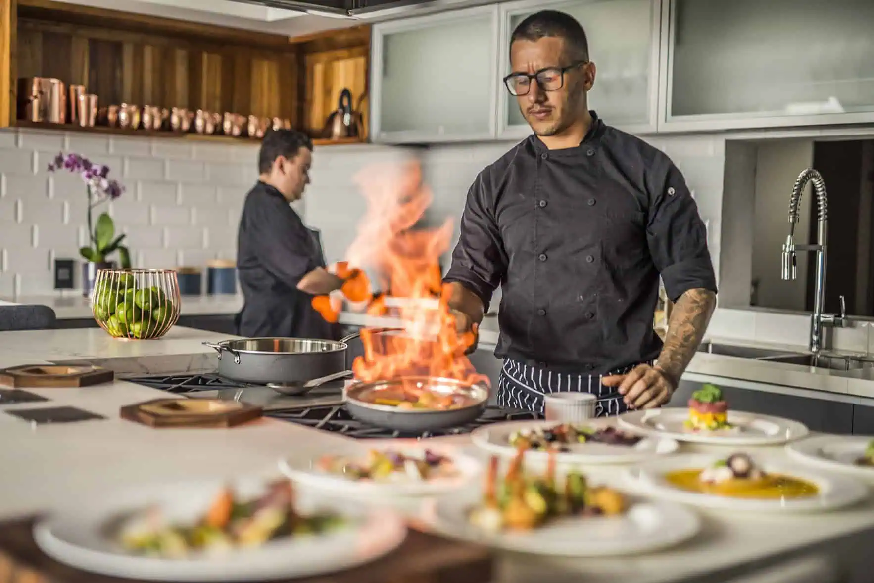 Professional chef flambeing food in a modern kitchen with an assistant chef in the background.