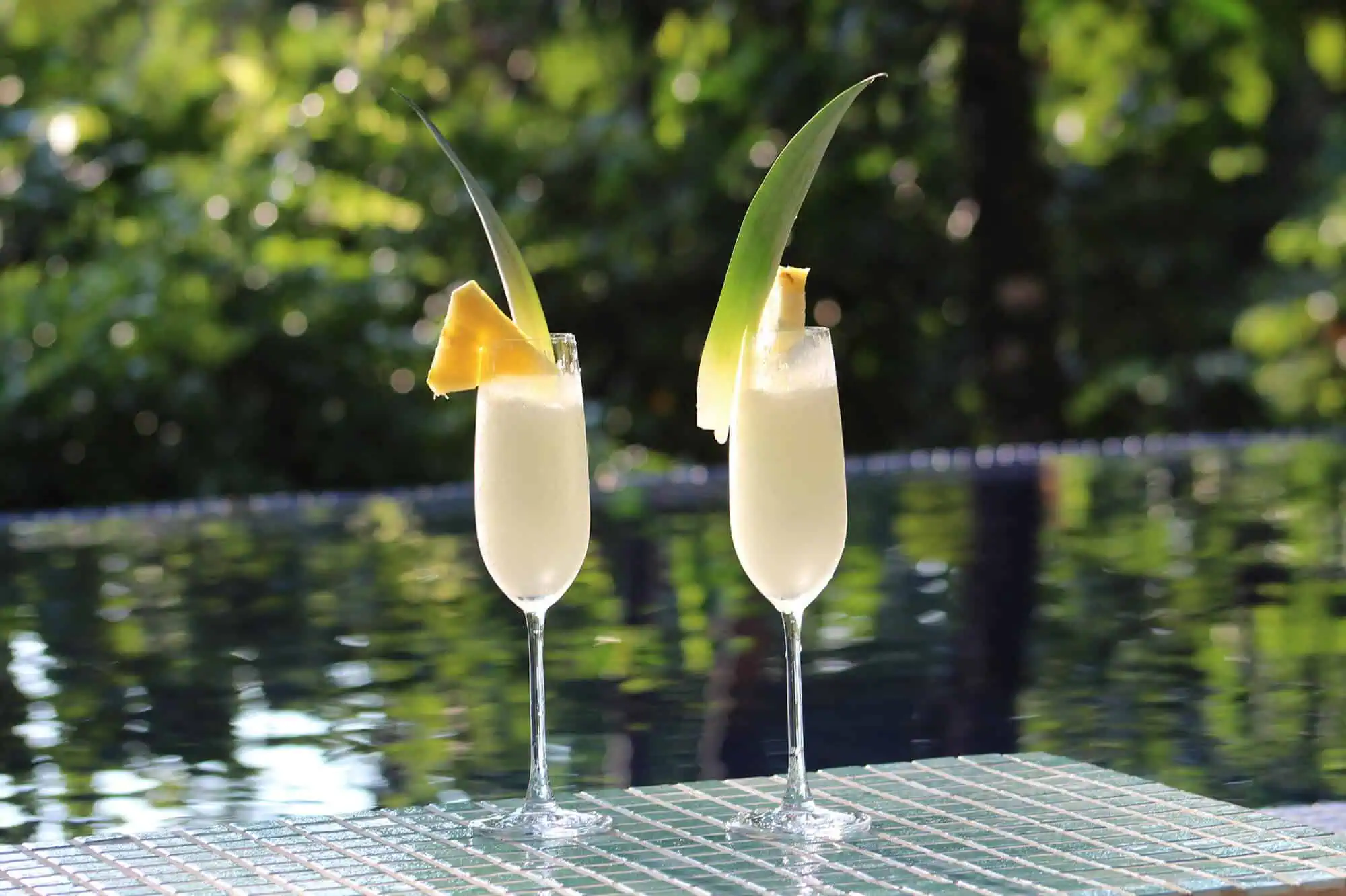 Two tropical cocktails garnished with pineapple slices and palm leaves, set on a mosaic table by a pool.