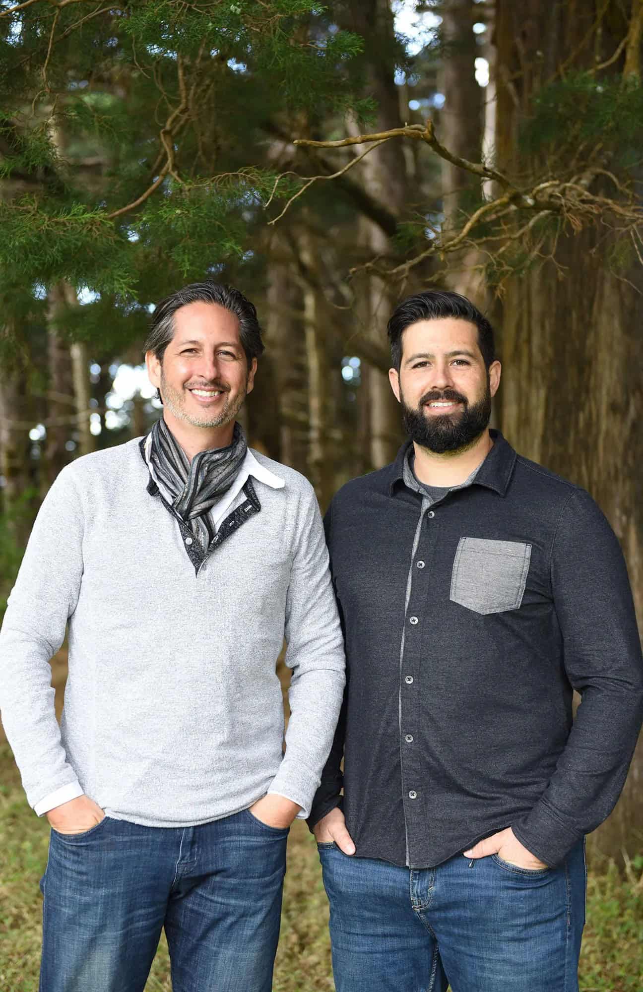 Two smiling men standing together outdoors with a backdrop of tall trees.