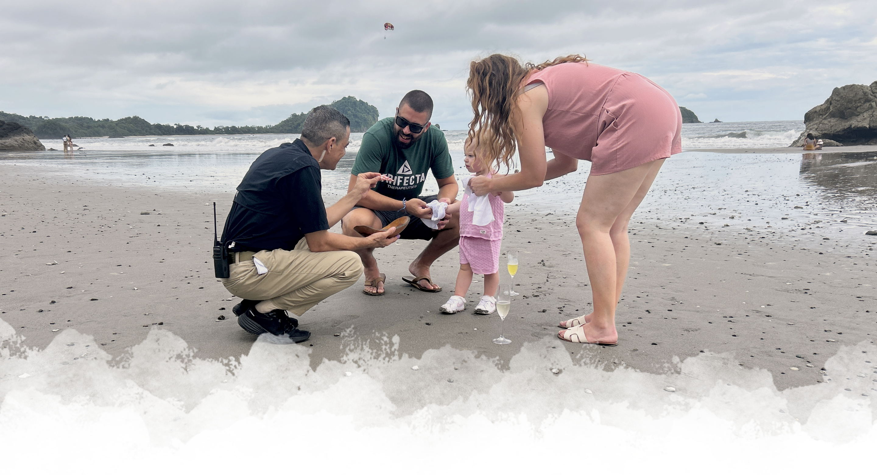 Family moment on a beach with a man squatting and showing something to another man, while a woman helps a young child with her toy, with waves and a rocky island in the background