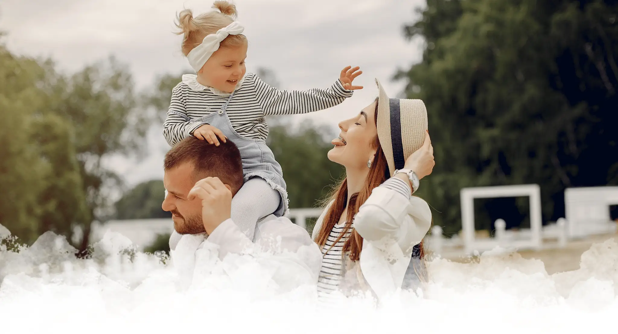 Joyful family moment outdoors with a young child gleefully sitting atop her father's shoulders, reaching out to her smiling mother who wears a stylish straw hat, surrounded by lush greenery.