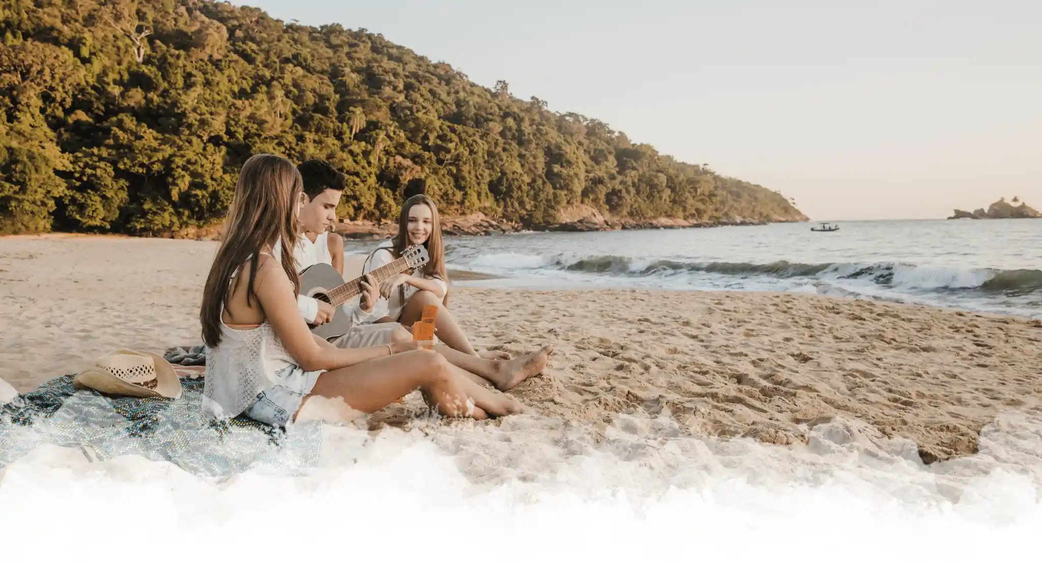 two women and one man sitting on a blanket. The man plays guitar while one woman sings along, against a backdrop of gentle waves and a lush green hillside