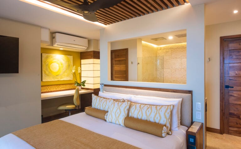 Elegantly designed bedroom featuring a cozy bed with stylish golden pillows, an artistic yellow painting above a desk space, and a visible luxurious bathroom area, all framed by wooden accents and warm ambient lighting.