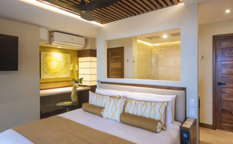 Elegantly designed bedroom featuring a cozy bed with stylish golden pillows, an artistic yellow painting above a desk space, and a visible luxurious bathroom area, all framed by wooden accents and warm ambient lighting.