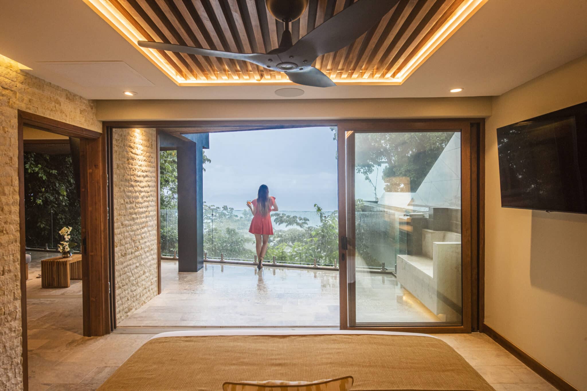 Spacious and elegant hotel room with a breathtaking ocean view, featuring a woman in a pink dress enjoying the scenery from a large open balcony. The room is well-lit with natural light, accentuated by stylish wood and stone architecture and modern decor