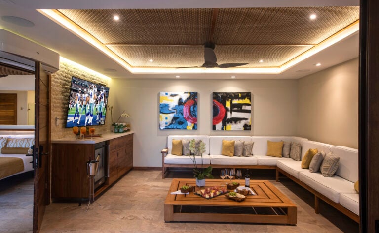 Contemporary entertainment lounge with a large flat-screen TV displaying a sports game, flanked by vibrant abstract paintings and a long white sectional sofa with golden cushions, set against a backdrop of rich wooden cabinetry and modern lighting.