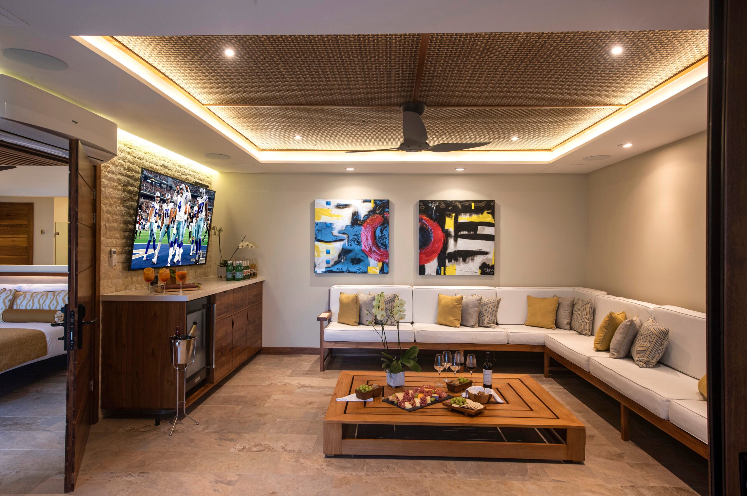 Contemporary entertainment lounge with a large flat-screen TV displaying a sports game, flanked by vibrant abstract paintings and a long white sectional sofa with golden cushions, set against a backdrop of rich wooden cabinetry and modern lighting.