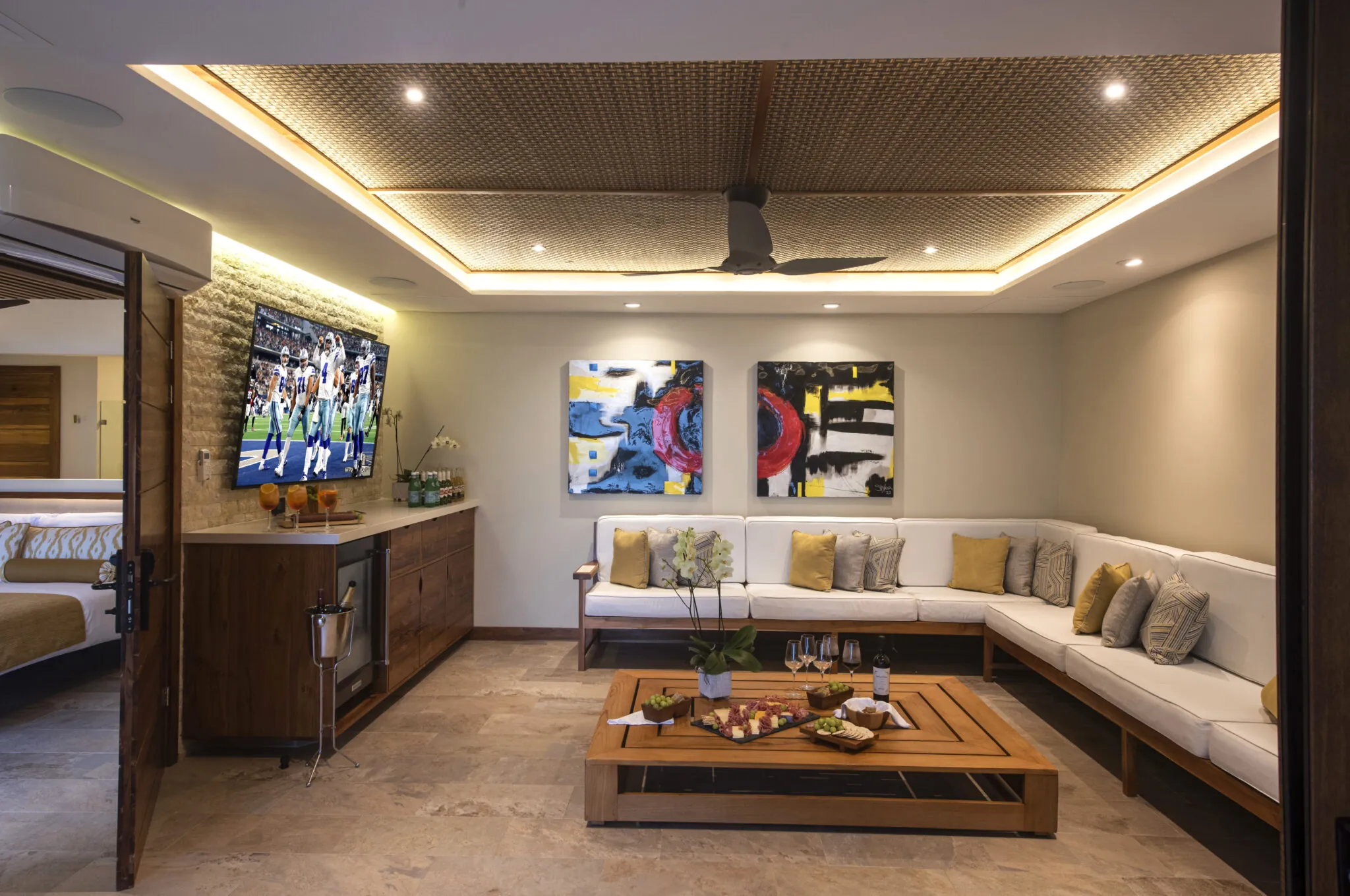 Contemporary entertainment lounge with a large flat-screen TV displaying a sports game, flanked by vibrant abstract paintings and a long white sectional sofa with golden cushions, set against a backdrop of rich wooden cabinetry and modern lighting.
