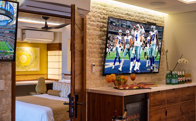 Luxury suite with a spacious layout featuring a large flat-screen TV showing a football game, connected to a cozy bedroom with elegant decor. The room includes a well-stocked minibar and decorative elements, offering a view through the open door.