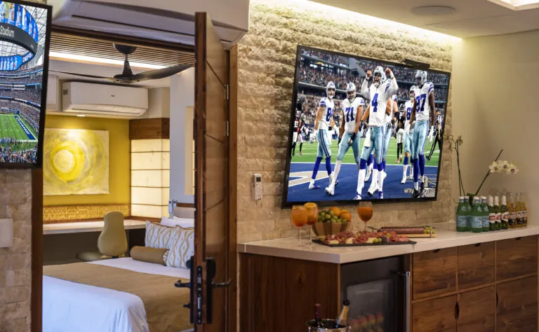 Luxury suite with a spacious layout featuring a large flat-screen TV showing a football game, connected to a cozy bedroom with elegant decor. The room includes a well-stocked minibar and decorative elements, offering a view through the open door.