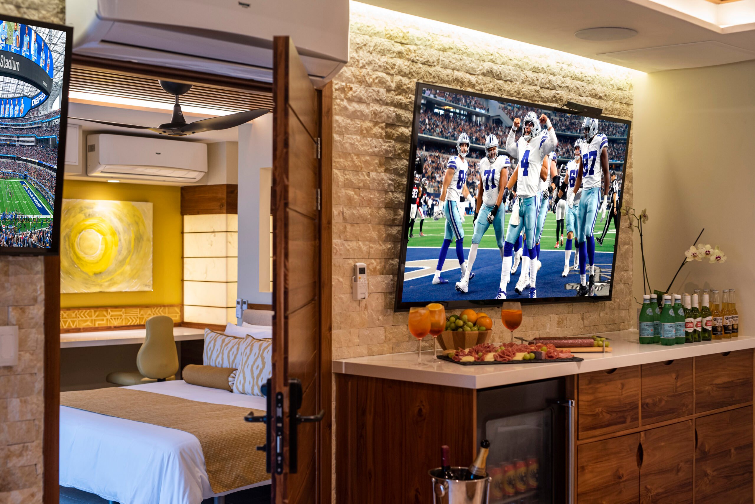 Luxury suite with a spacious layout featuring a large flat-screen TV showing a football game, connected to a cozy bedroom with elegant decor. The room includes a well-stocked minibar and decorative elements, offering a view through the open door.