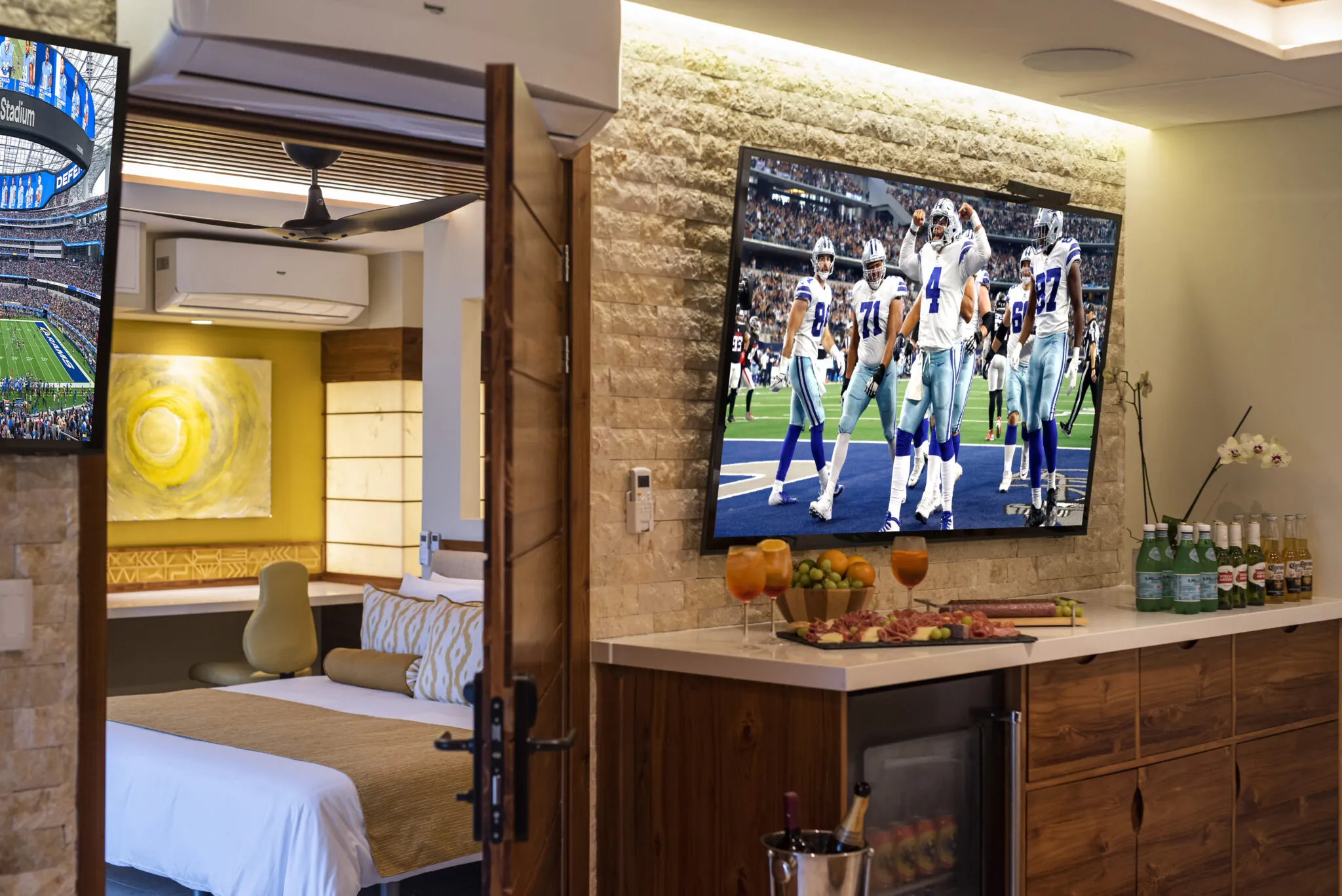 Luxury suite with a spacious layout featuring a large flat-screen TV showing a football game, connected to a cozy bedroom with elegant decor. The room includes a well-stocked minibar and decorative elements, offering a view through the open door.
